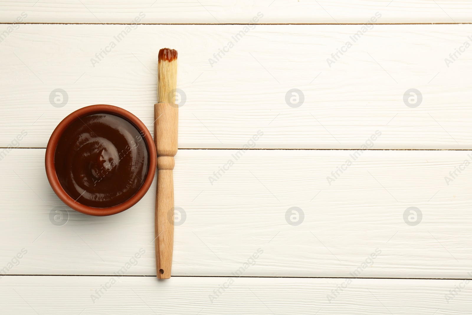 Photo of Tasty barbeque sauce in bowl and brush on white wooden table, top view. Space for text