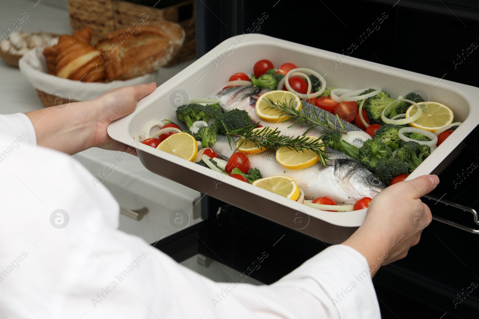 Photo of Woman putting baking dish with raw fish and vegetables into oven in kitchen, closeup