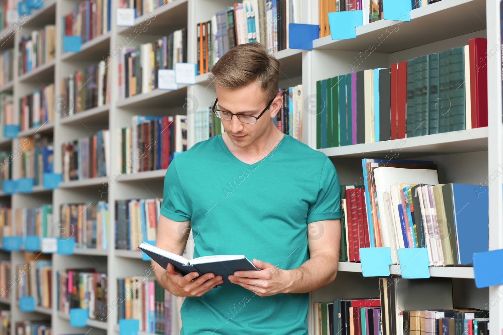 Photo of Young man with book near shelving unit in library