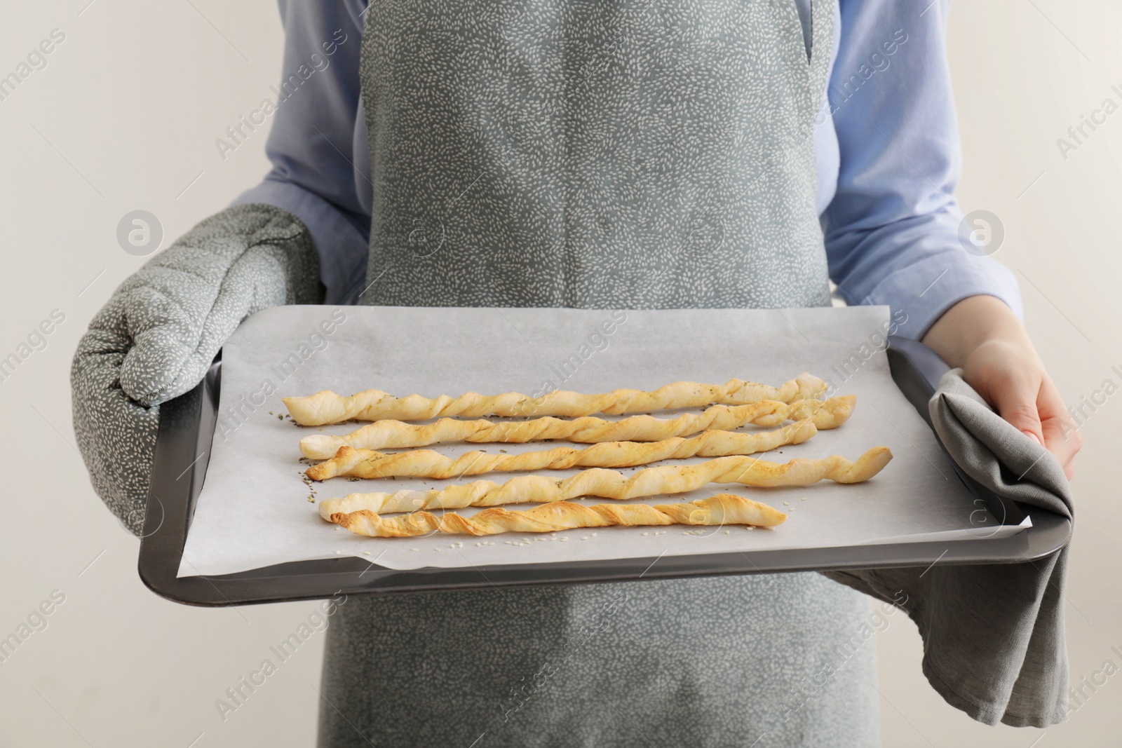 Photo of Woman holding baking sheet with homemade breadsticks on light grey background, closeup. Cooking traditional grissini