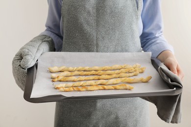 Photo of Woman holding baking sheet with homemade breadsticks on light grey background, closeup. Cooking traditional grissini