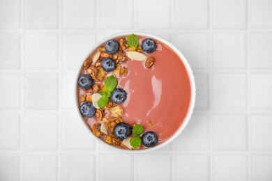 Bowl of delicious smoothie with fresh blueberries and granola on white tiled table, top view