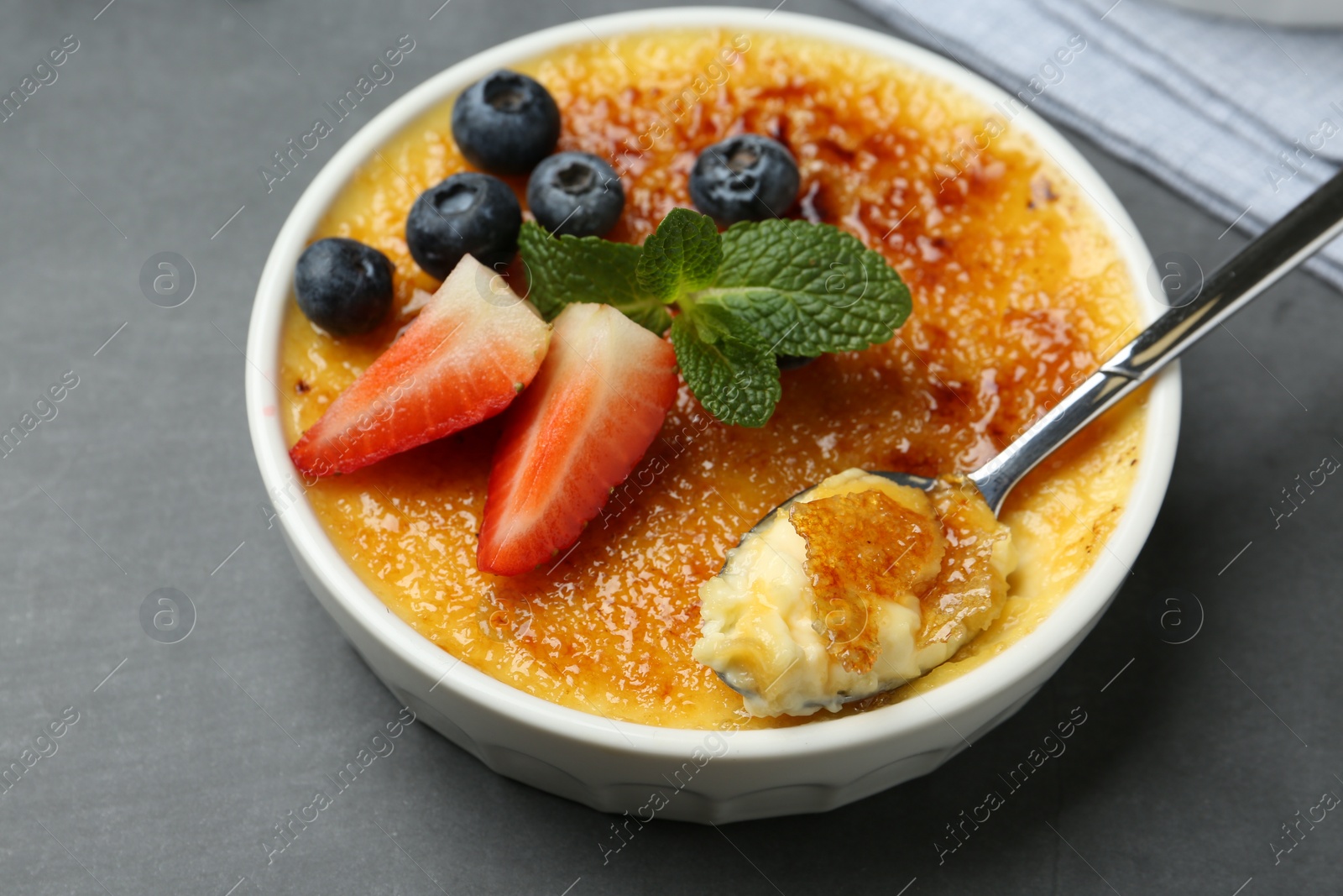 Photo of Delicious creme brulee with berries in bowl and spoon on grey table, closeup