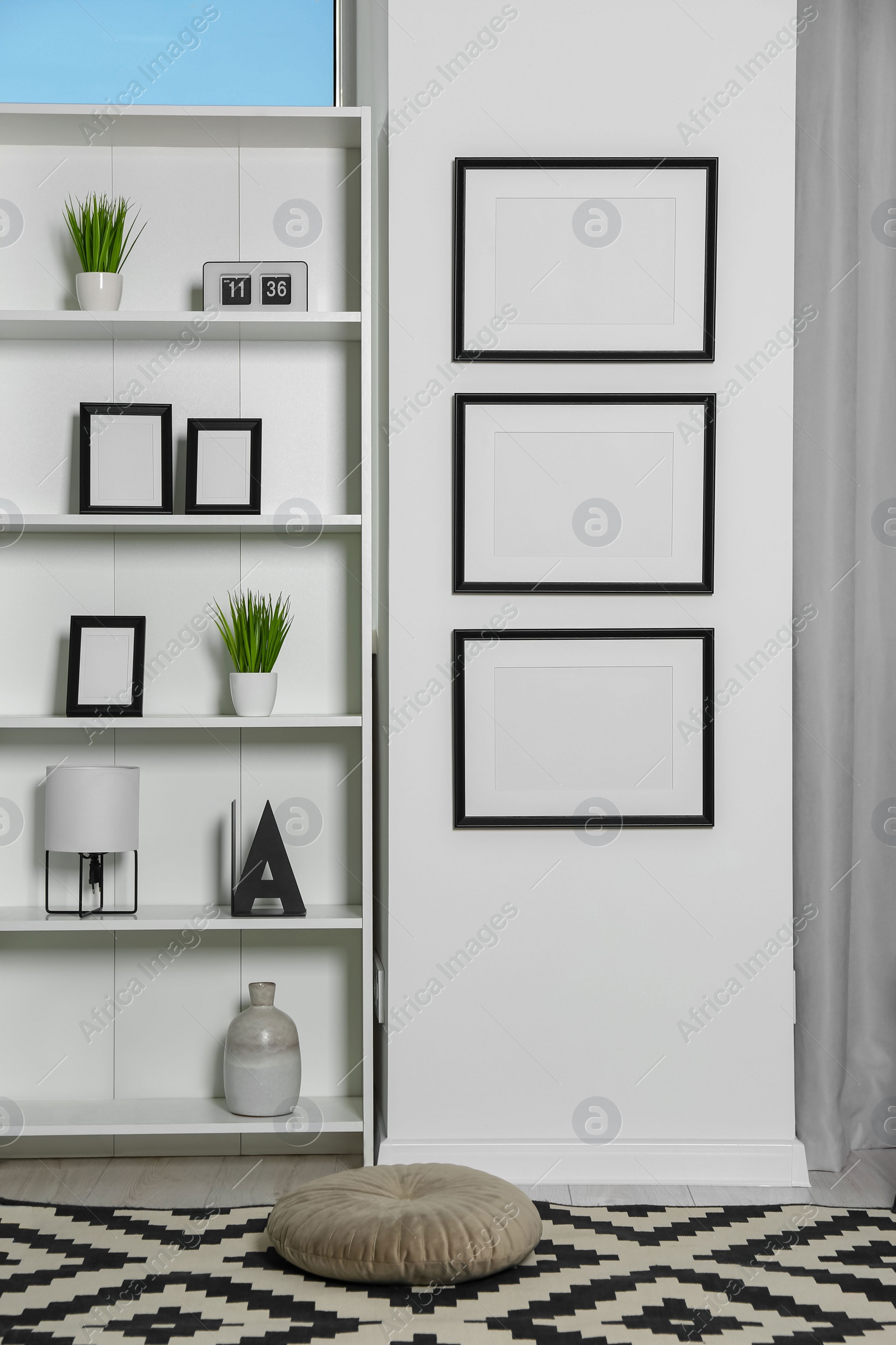 Photo of Stylish room interior with empty frames hanging on white wall near shelving unit