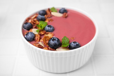 Photo of Bowl of delicious smoothie with fresh blueberries and granola on white tiled table, closeup