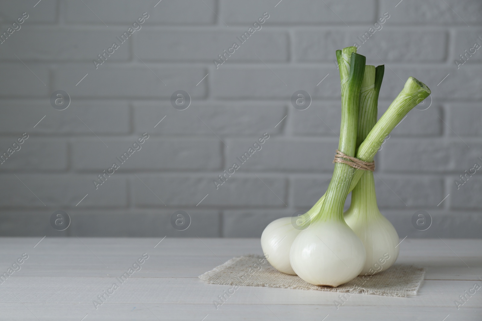 Photo of Bunch of fresh green spring onions on white wooden table against brick wall, space for text