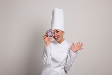 Photo of Happy professional confectioner in uniform holding delicious doughnut on light grey background