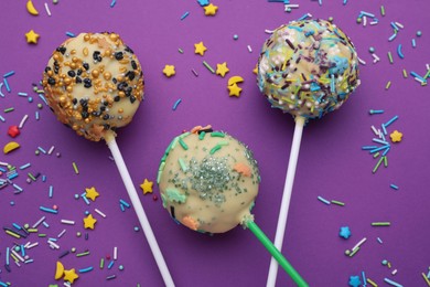 Photo of Sweet cake pops and sprinkles on purple background, flat lay. Delicious confectionery
