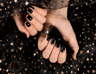 Photo of Woman with black manicure holding nail polish bottle over dark fabric, top view