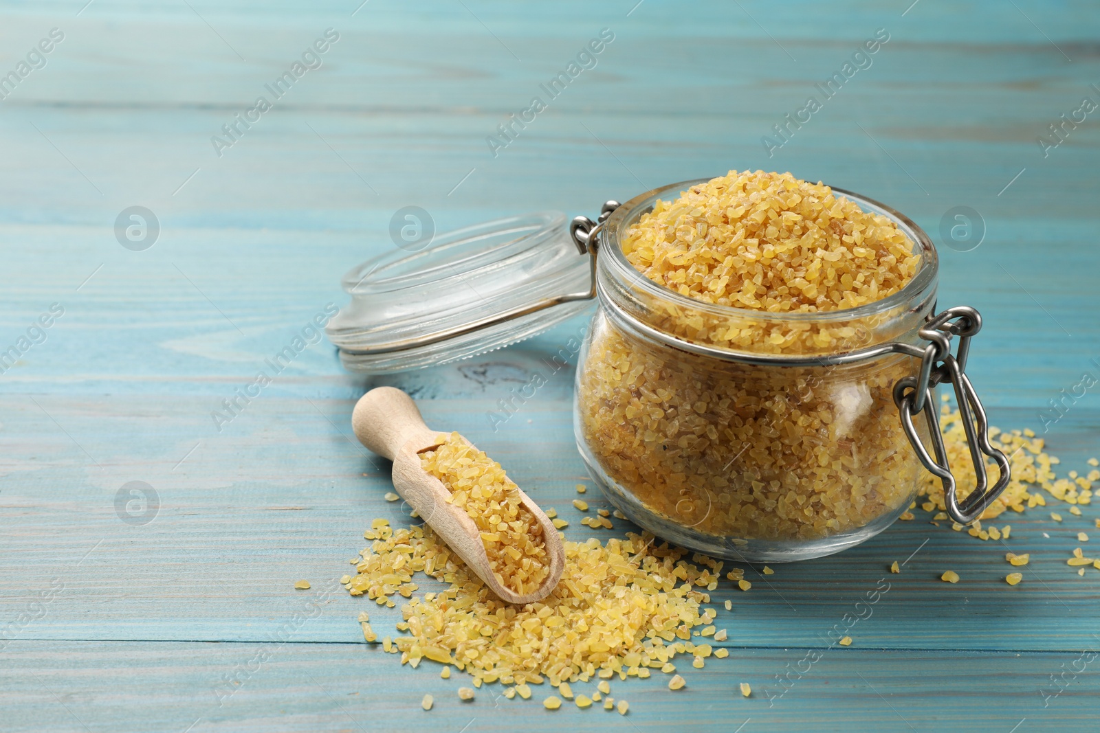 Photo of Glass jar and scoop with raw bulgur on light blue wooden table