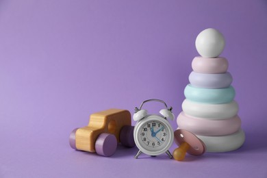 Alarm clock, toys and baby dummy on violet background, space for text. Time to give birth