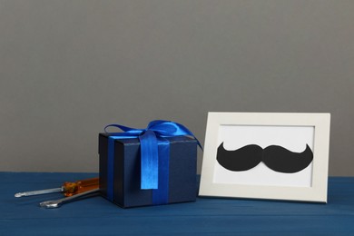 Photo of Happy Father's Day. Gift box, tools and frame with mustache on blue wooden table