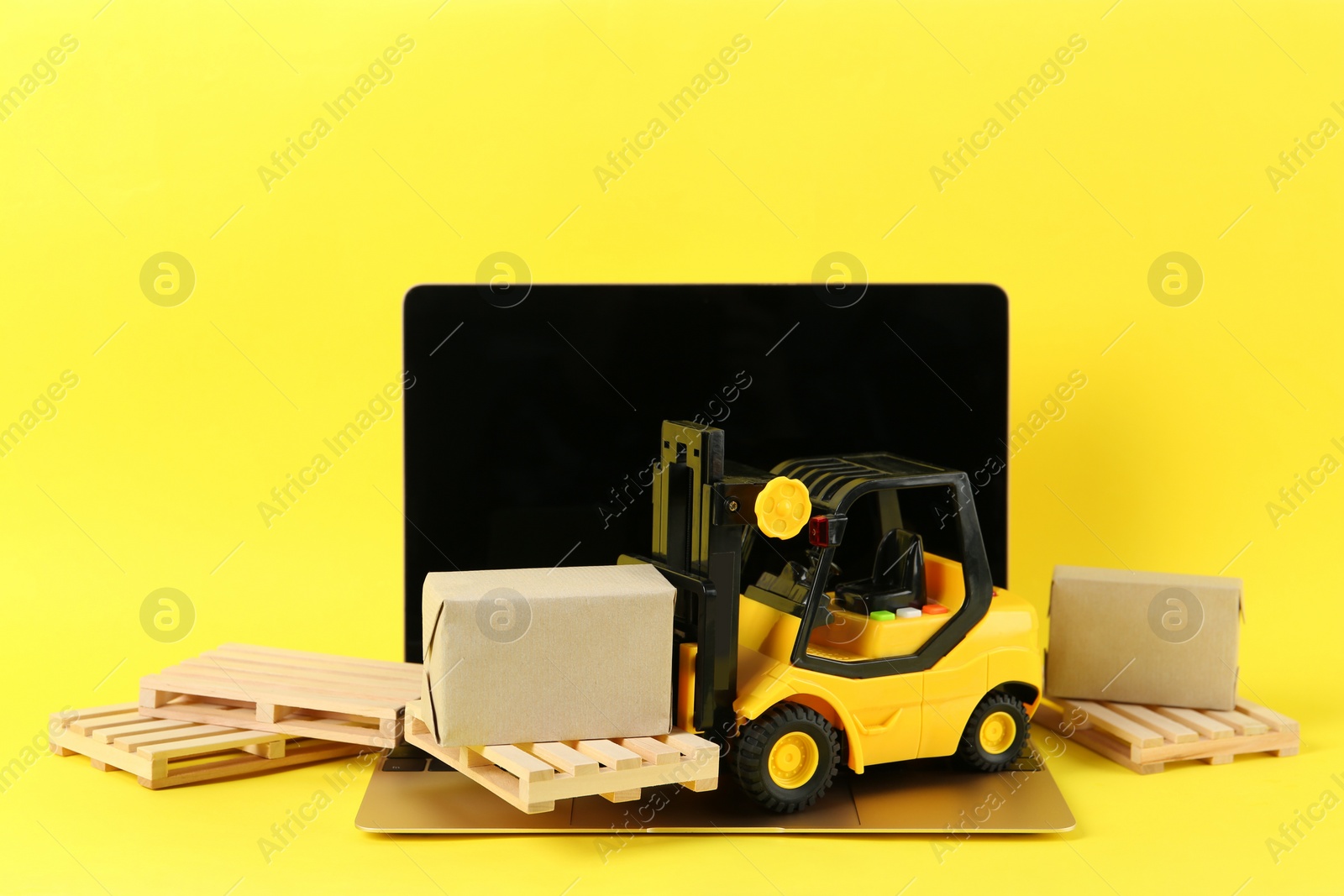 Photo of Laptop, toy forklift with wooden pallets and boxes on yellow background
