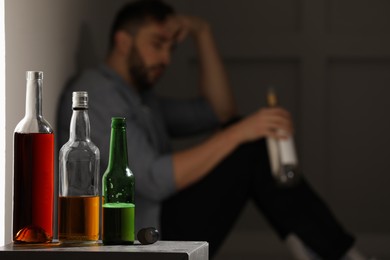 Addicted man sitting near wall indoors, focus on table with alcoholic drinks. Space for text