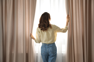 Woman opening window curtains at home in morning, back view