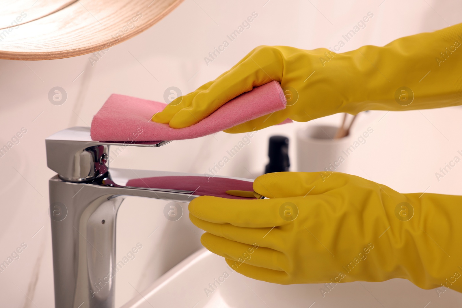 Photo of Woman in gloves cleaning faucet of bathroom sink with rag, closeup