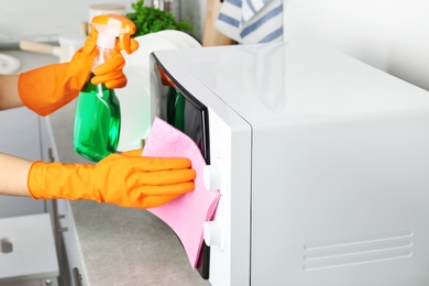 Photo of Woman cleaning microwave oven with rag and detergent in kitchen, closeup