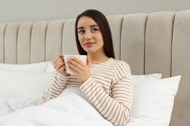 Beautiful young woman with cup of drink in bed at home