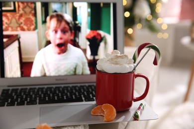 MYKOLAIV, UKRAINE - DECEMBER 25, 2020: Laptop displaying Home Alone movie indoors, focus on cup of sweet drink and tangerine slices. Cozy winter holidays atmosphere