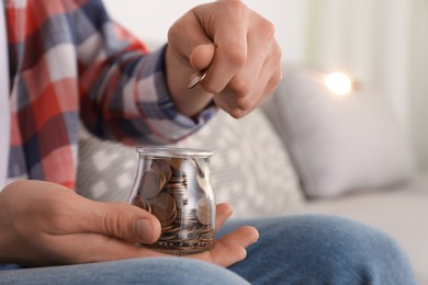 Photo of Man putting coin into glass jar at home, closeup