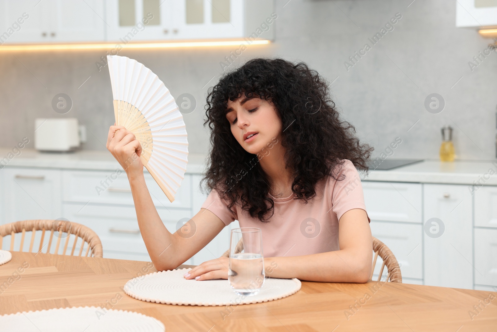 Photo of Young woman waving hand fan to cool herself at table in kitchen