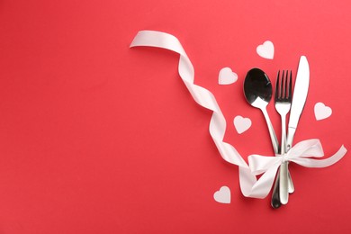 Beautiful cutlery set, hearts and white bow on red background, flat lay with space for text. Valentine's Day dinner