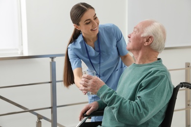 Photo of Nurse giving water to senior man in wheelchair at hospital. Medical assisting