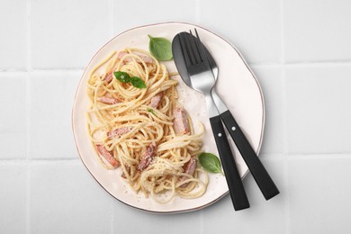 Photo of Plate of tasty pasta Carbonara with basil leaves, fork and spoon on white tiled table, top view