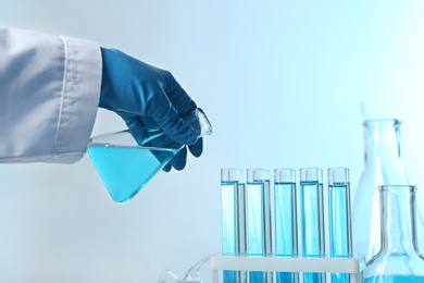 Scientist pouring color liquid from conical flask into test tubes on light background, closeup with space for text. Solution chemistry