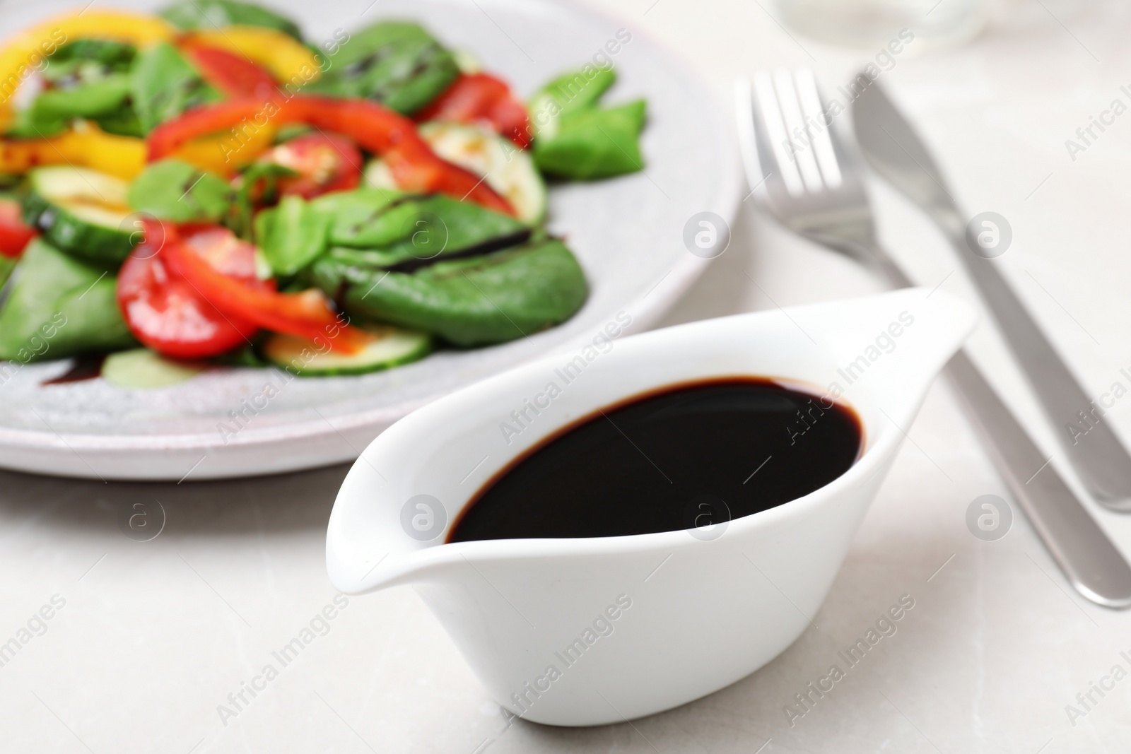 Photo of Balsamic vinegar in gravy boat near plate with vegetable salad on table, closeup