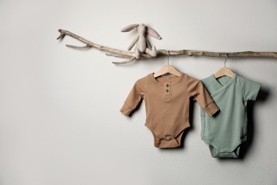 Photo of Baby bodysuits and toy on decorative branch near light wall. Space for text