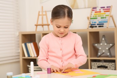 Photo of Cute little girl using colorful glitter while making paper card at desk in room. Home workplace