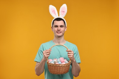 Easter celebration. Handsome young man with bunny ears holding basket of painted eggs on orange background