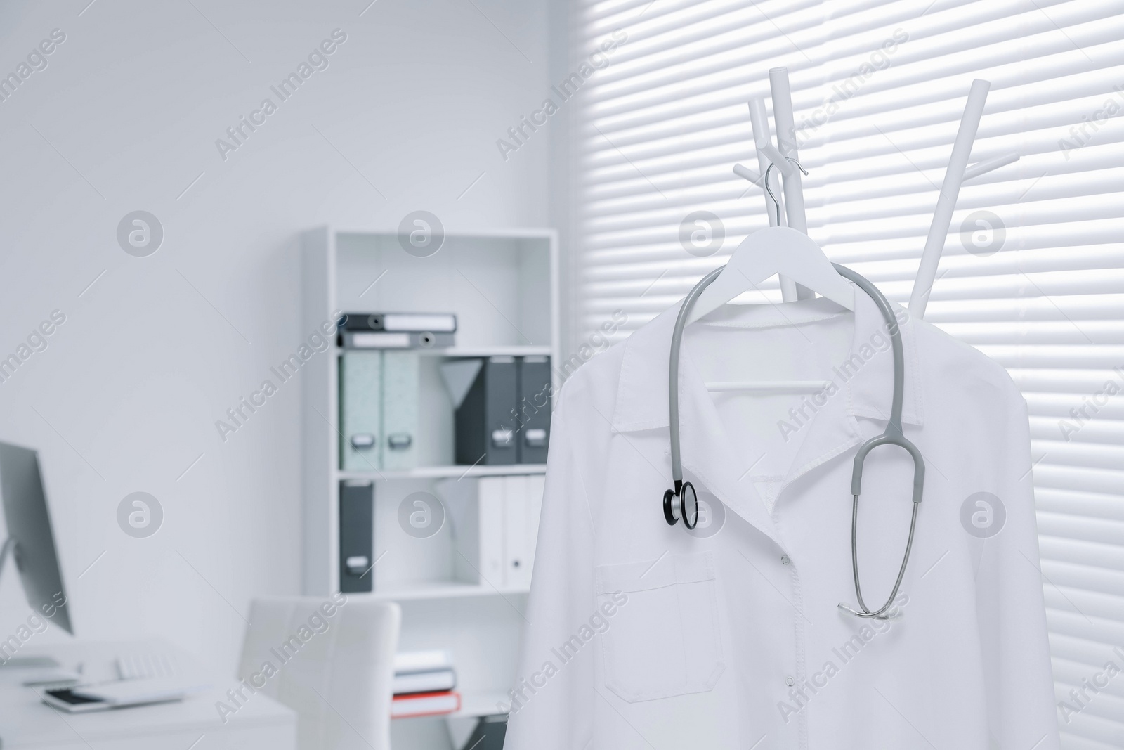 Photo of White doctor's gown and stethoscope hanging on rack in clinic, closeup. Space for text