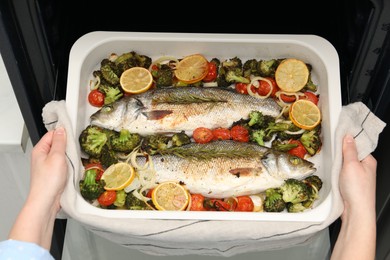 Photo of Woman taking baking dish with delicious fish and vegetables from oven, top view