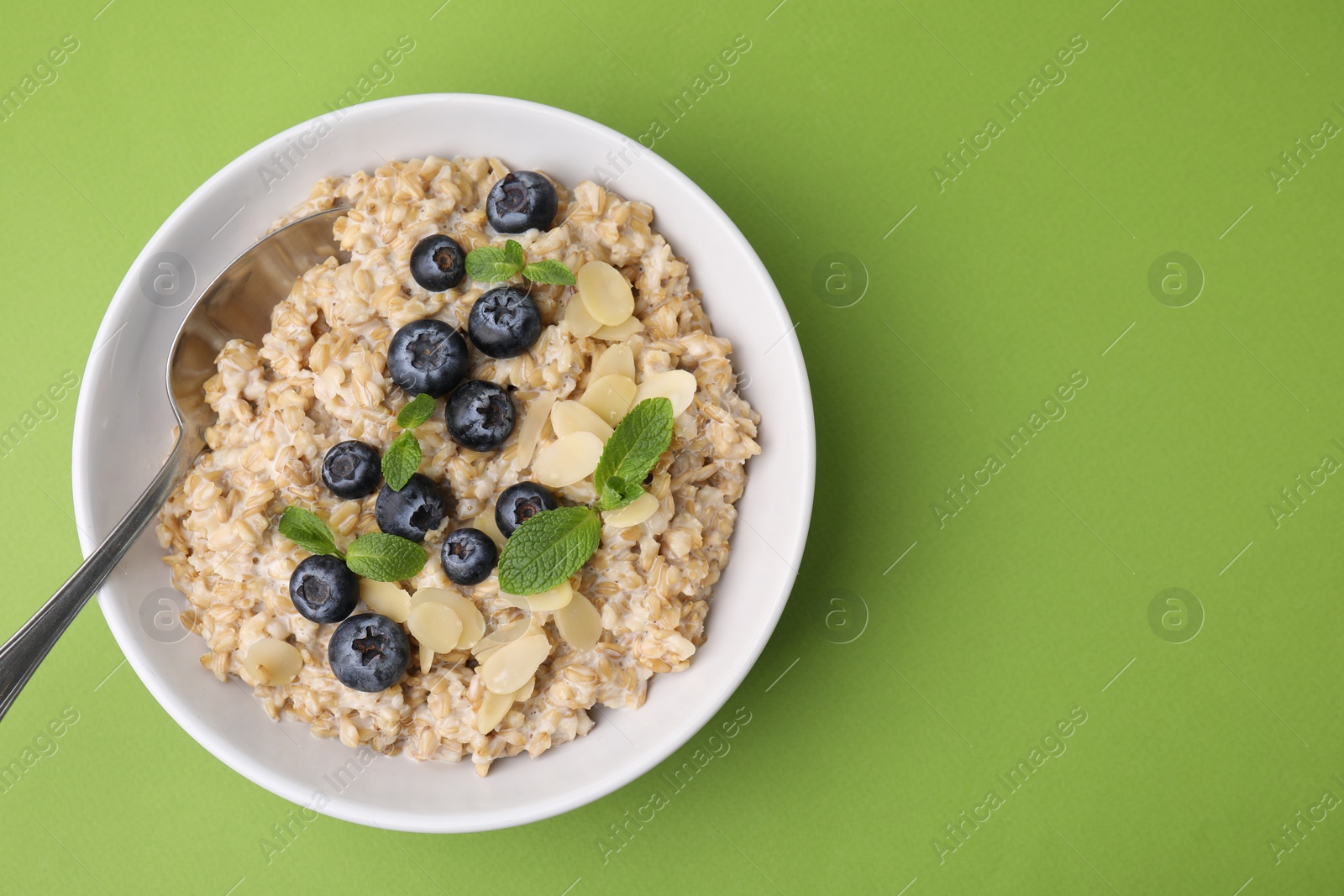 Photo of Tasty oatmeal with blueberries, mint and almond petals in bowl on light green background, top view. Space for text