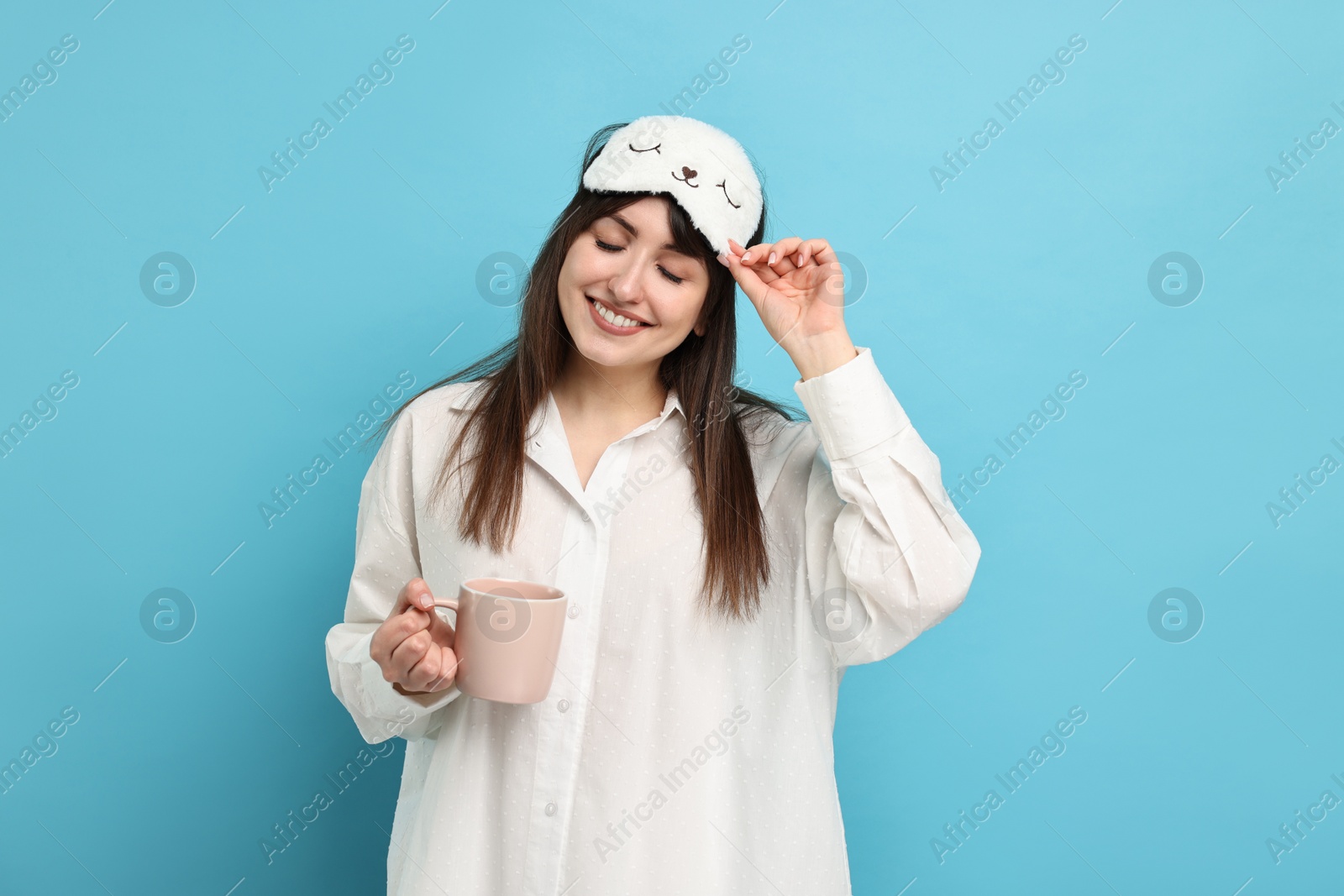 Photo of Woman in pyjama and sleep mask holding cup of drink on light blue background