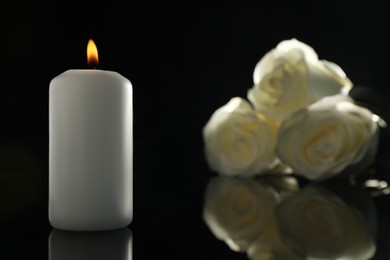 White roses and burning candle on black mirror surface in darkness, closeup with space for text. Funeral symbols