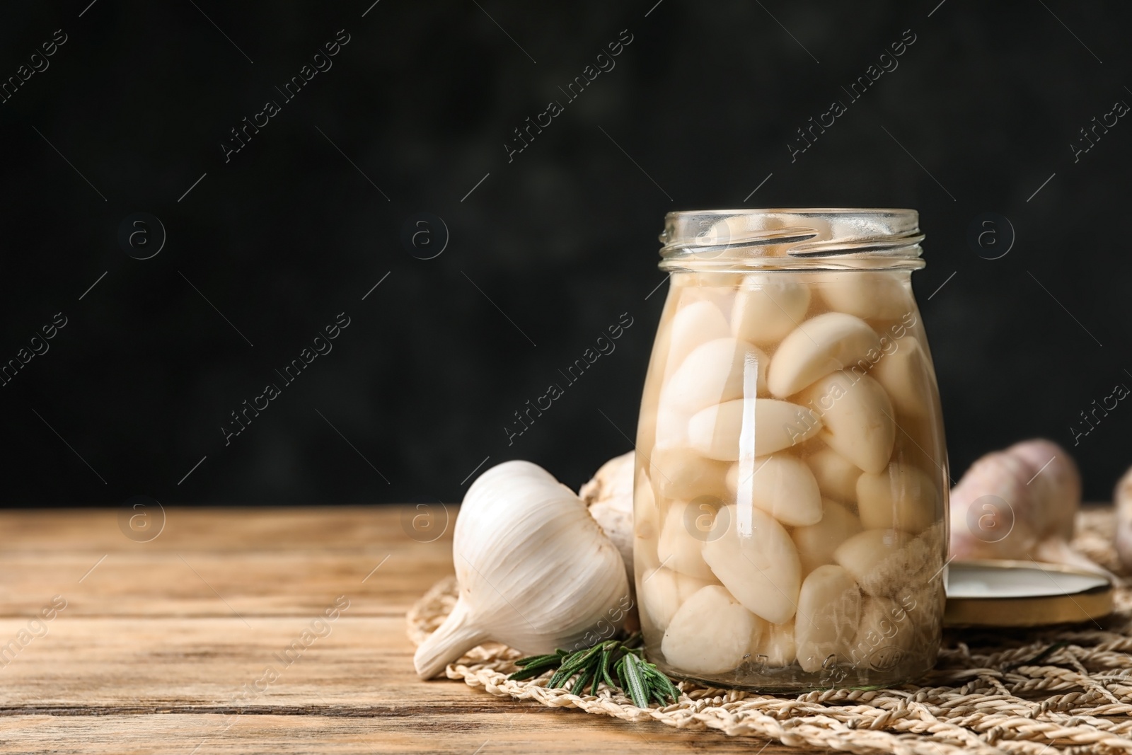 Photo of Preserved garlic in glass jar on wooden table against dark background. Space for text