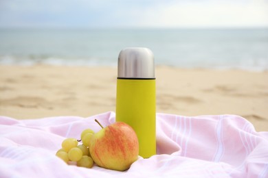 Metallic thermos with hot drink, fruits and plaid on sandy beach near sea