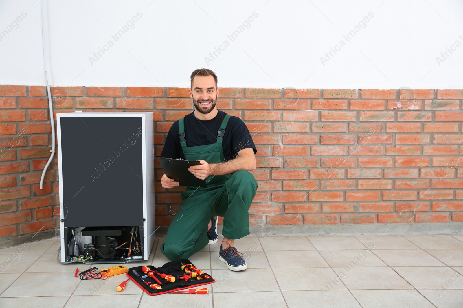 Photo of Male technician with clipboard and tools near broken refrigerator indoors. Space for text
