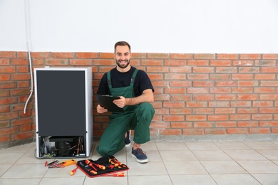 Photo of Male technician with clipboard and tools near broken refrigerator indoors. Space for text