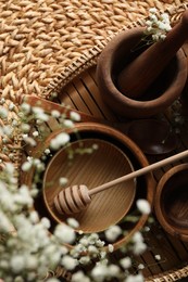 Wooden bowls, rolling pin and honey dipper near flowers on wicker mat, flat lay