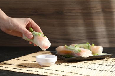 Photo of Woman dipping delicious spring roll into sauce at black table, closeup