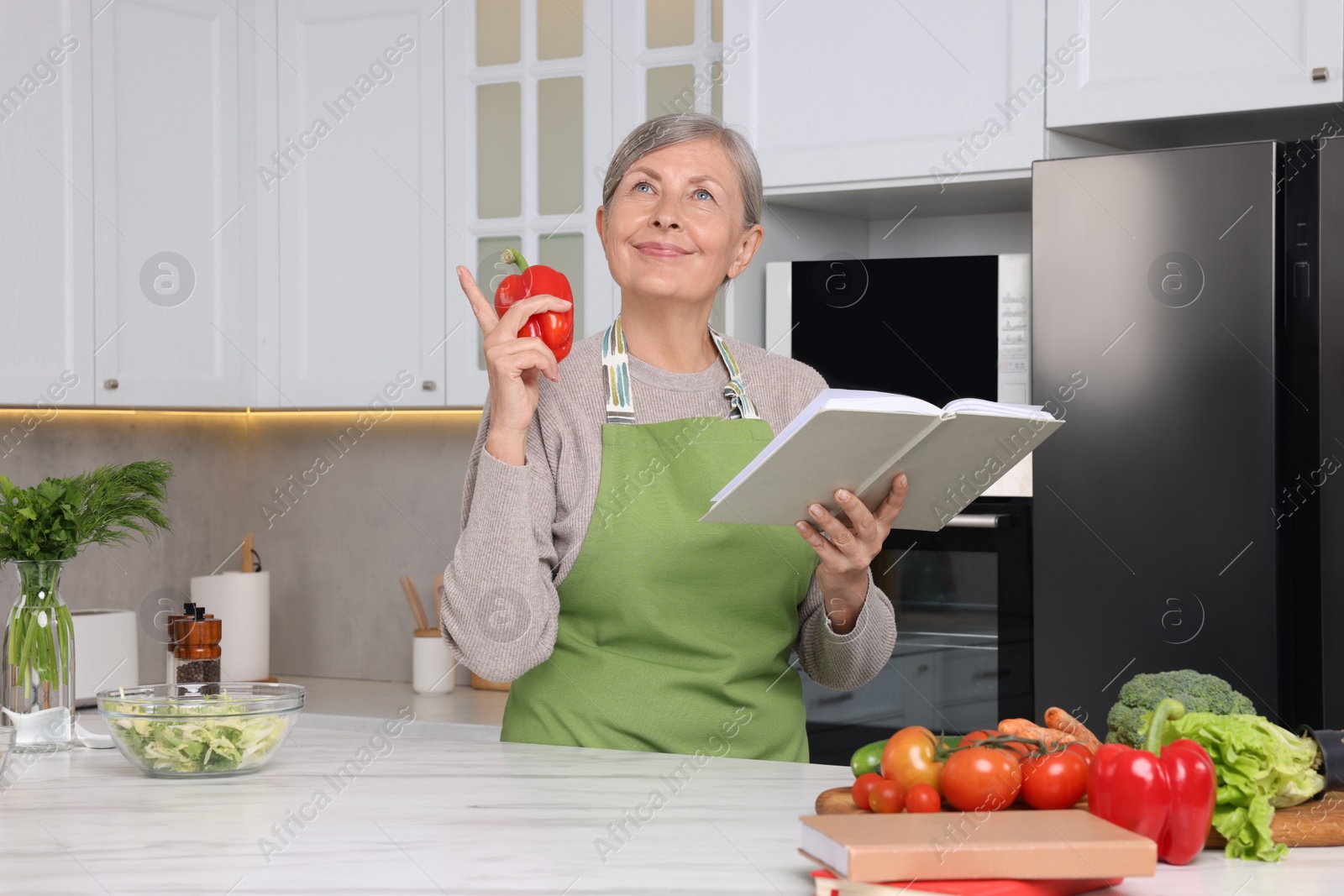 Photo of Happy woman with recipe book and bell pepper in kitchen