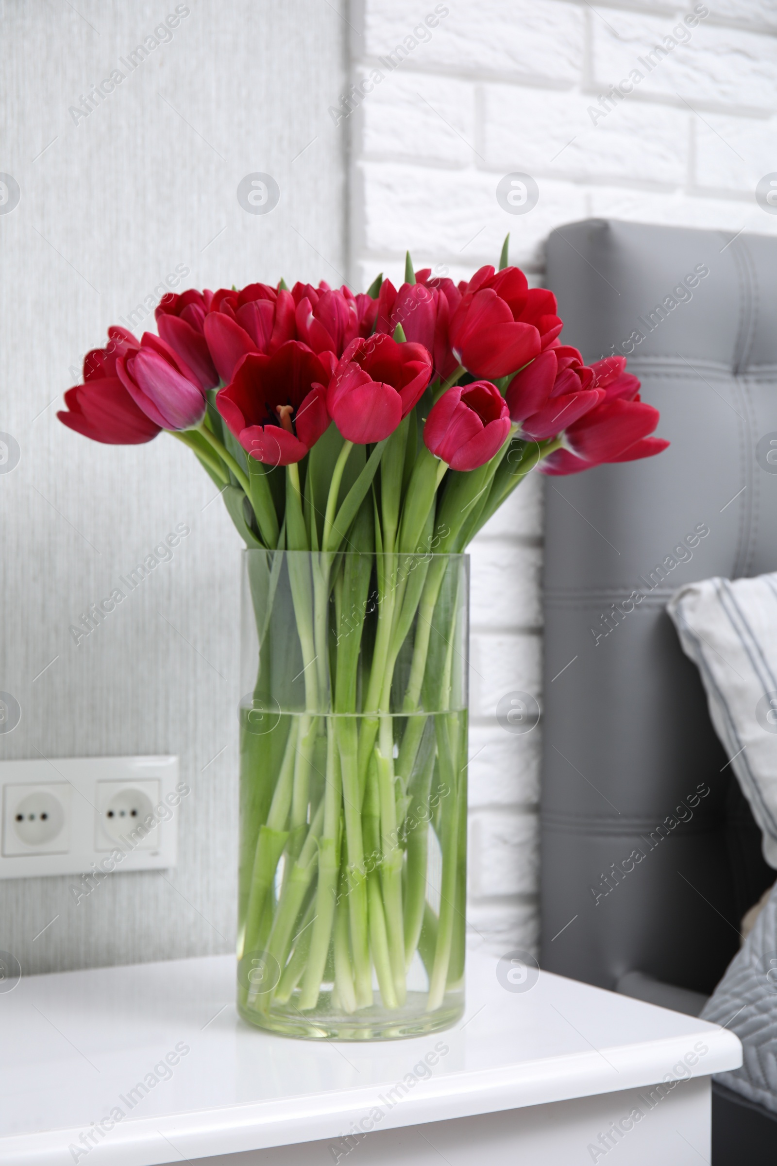 Photo of Bouquet of beautiful tulips in glass vase on white nightstand near bed