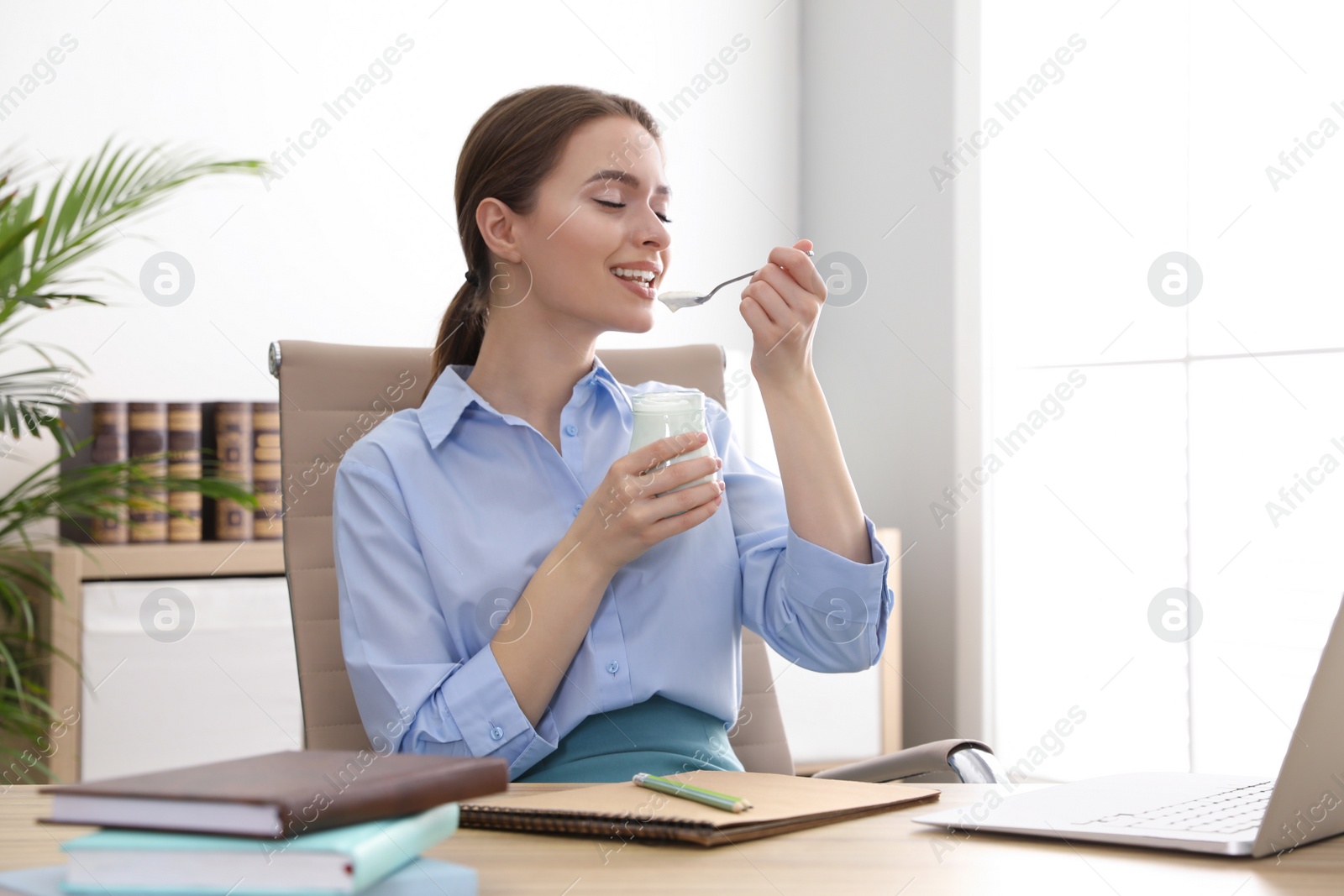 Photo of Young attractive woman eating tasty yogurt at table in office