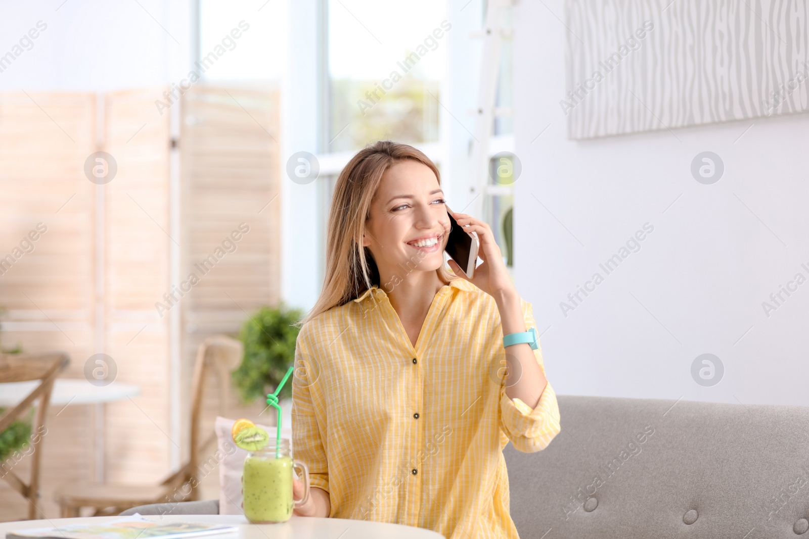 Photo of Young woman using mobile phone while drinking tasty healthy smoothie at table, indoors