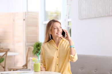 Young woman using mobile phone while drinking tasty healthy smoothie at table, indoors