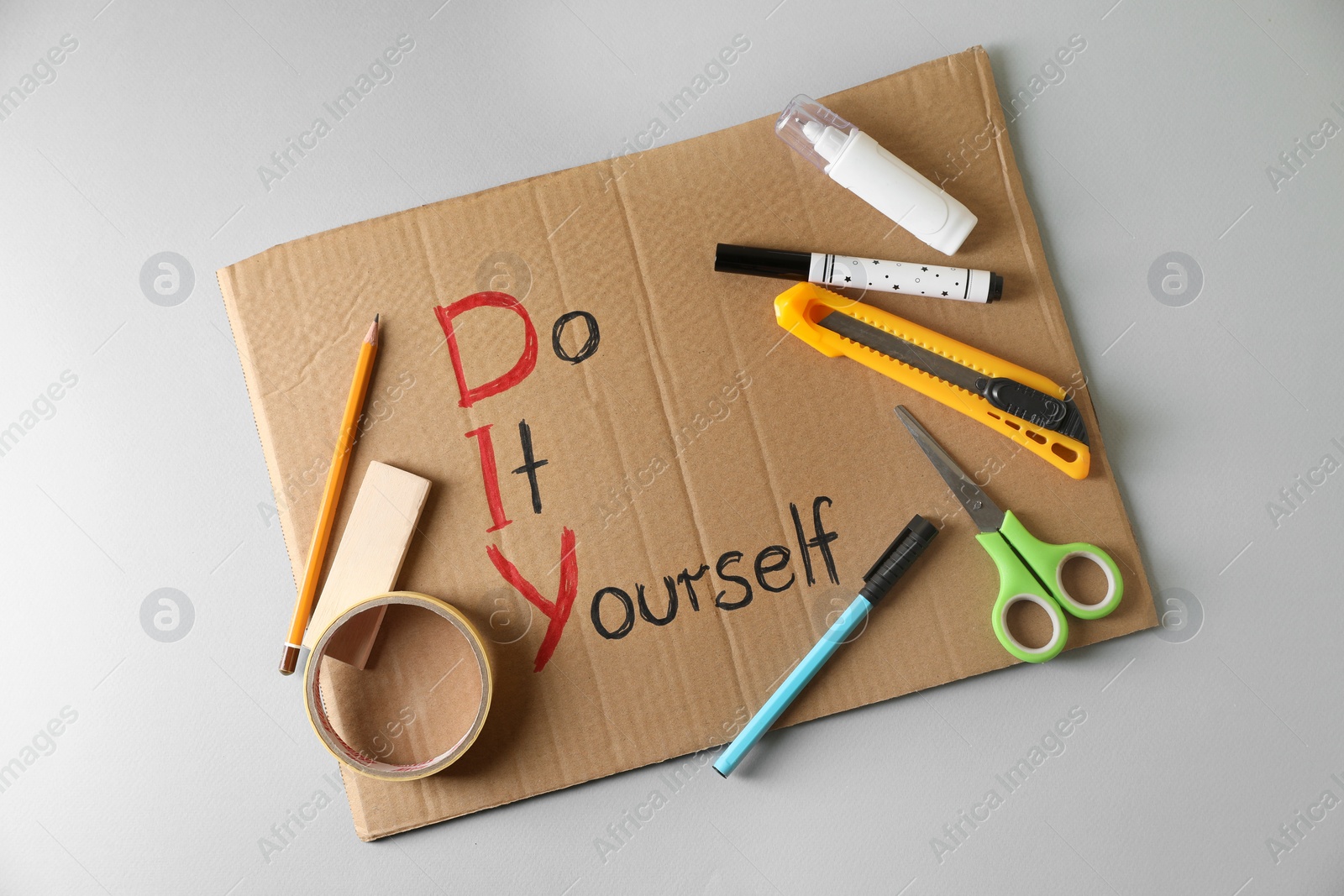 Photo of Cardboard sheet with written phrase Do It Yourself and stationery on light grey background, top view. DIY concept